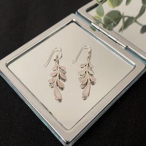 Può includere: Un paio di orecchini pendenti color argento con un design a forma di foglia. Gli orecchini sono esposti su una superficie a specchio, che riflette gli intricati dettagli dei gioielli. Chiusura a gancio.