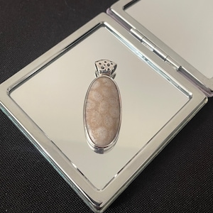 May include: A silver pendant with a light brown, oval-shaped stone. The pendant is set on a silver frame and rests on a mirrored surface. The pendant has a decorative top with small cut-out details. The background is black.