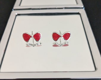 May include: A pair of bow-shaped stud earrings. Each earring features a bright red, textured center framed by a silver-colored metal. The earrings are displayed on a reflective surface, showing their mirrored image.