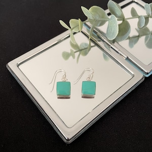 May include: A pair of silver drop earrings featuring square-shaped, smooth turquoise-coloured stones. Each earring is suspended from a simple silver hook wire, showcasing a clean, geometric design that reflects clearly on the mirrored surface.