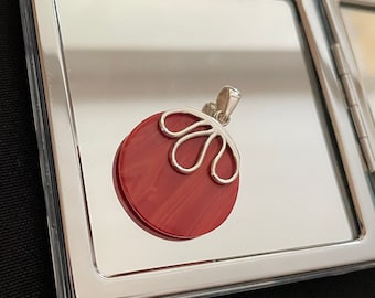 Puede incluir: Un colgante de coral rojo con un diseño de filigrana de plata. El colgante es redondo y tiene una superficie lisa y pulida.