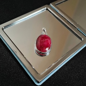Puede incluir: Un colgante de plata con una piedra preciosa roja. El colgante tiene forma ovalada y un acabado liso y pulido. La piedra preciosa es de color rojo intenso y tiene un ligero brillo.