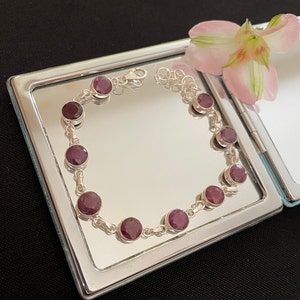 Può includere: Un braccialetto d'argento con un design a catena e otto pietre rotonde di colore rosso scuro. Il braccialetto è esposto su un vassoio d'argento con una superficie a specchio.