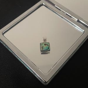 Puede incluir: Un colgante de plata con una incrustación cuadrada de nácar de abulón. La concha tiene un brillo iridiscente verde, azul y marrón.
