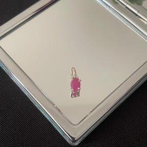 May include: A small, silver pendant with a vibrant, oval-shaped ruby gemstone. The ruby is a deep pinkish-red color and is set in a silver metal setting. The pendant is displayed on a reflective surface.
