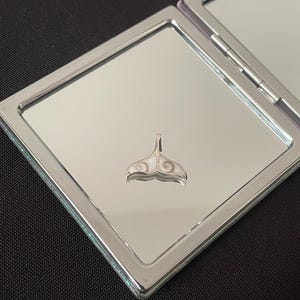 Peut inclure: Un miroir compact argenté avec un charme de queue de baleine blanche et brune. Le miroir est fermé et le charme est au centre du miroir.