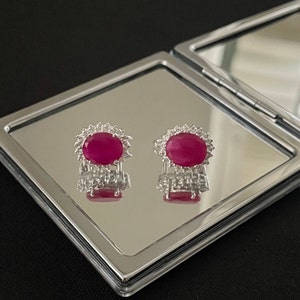 Op de afbeelding: Een paar robijn- en diamanten oorbellen, gepresenteerd op een compacte spiegel. Elke oorbel heeft een grote, ovaal geslepen robijn omringd door kleine diamanten. De oorbellen zijn gezet in een zilveren of witgouden zetting.