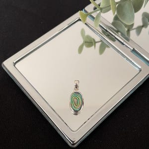 Könnte beinhalten: Ein ovaler Anhänger mit einer Einlage aus poliertem Abalone-Muschelmaterial in schillernden Wirbeln aus Grün-, Blau- und Rosatönen, eingefasst in einen glatten, silberfarbenen Metallrahmen mit einer kleinen Öse an der Oberseite für eine Kette.