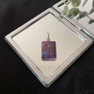 May include: A rectangular pendant with a silver bail, showcasing a mosaic of purple, blue, and gold tones. The pendant is presented on a mirrored surface within a silver-framed compact, against a black background.