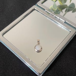 May include: A small, circular pendant featuring a smooth, white, opaque stone set within a polished silver-toned metal frame. The pendant includes a simple, triangular-shaped bail at the top for attachment to a chain or cord, reflecting light.