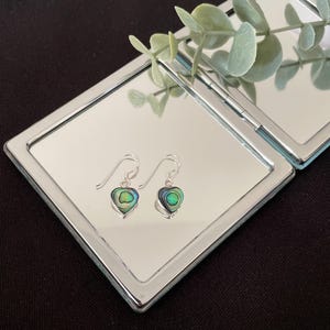 Può includere: Un paio di orecchini a gancio in argento con pendenti a forma di cuore intarsiati con conchiglia di abalone iridescente. Gli orecchini mostrano una vibrante miscela di toni verdi, blu e viola all'interno di una montatura in metallo argentato.