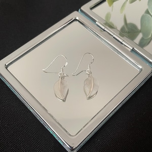 May include: A pair of silver-toned drop earrings featuring a leaf-shaped design with a central vein detail. The earrings are suspended from curved ear wires and are displayed on a reflective, square-shaped metallic surface with rounded corners.