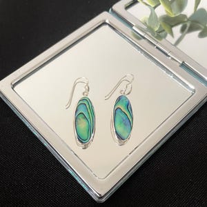 May include: A pair of oval-shaped drop earrings featuring iridescent abalone shell inlays set in polished silver-tone metal. Each earring hangs from a simple silver-tone hook, showcasing vibrant swirls of green, blue, and teal colours within the shell.