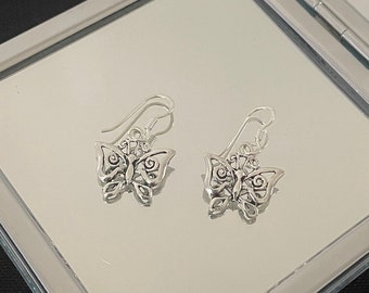 Könnte beinhalten: Ein Paar silberner Schmetterlingsohrringe mit filigranen Flügeldetails. Die Ohrringe werden auf einer reflektierenden Oberfläche präsentiert, die ihr zartes Design hervorhebt. Die Schmetterlingsanhänger hängen an einfachen Ohrhaken und wirken klassisch und elegant.