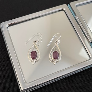 May include: A pair of silver dangle earrings with a teardrop-shaped design. Each earring features a round, purple gemstone.