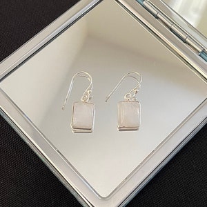 Puede incluir: Un par de pendientes de plata con piedras rectangulares blancas. Los pendientes están colgados de un espejo.