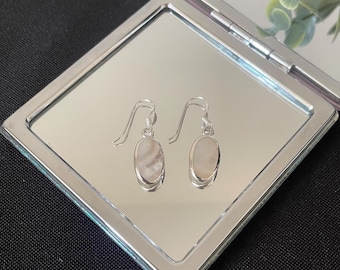 May include: A pair of silver dangle earrings with oval, light-colored stones. The earrings are displayed on a silver compact mirror, reflecting the earrings and the surrounding environment. The background is black.