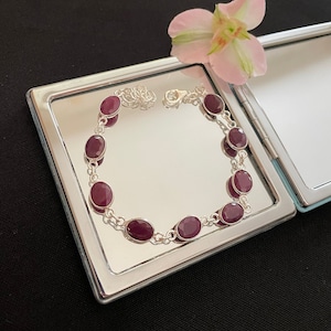 Puede incluir: Un brazalete de plata con una cadena y siete piedras ovaladas de color rojo oscuro. El brazalete se muestra sobre una superficie de espejo de plata.