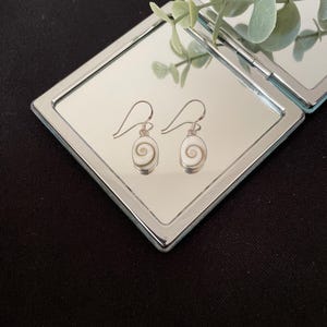 May include: A pair of drop earrings featuring oval-shaped white and tan Shiva eye shells set in silver-toned metal. Each shell displays a natural spiral pattern on the front, suspended from simple silver-toned ear wires for a clean look.