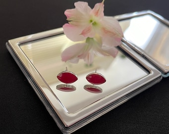 May include: A pair of silver earrings with red oval-shaped stones. The earrings are displayed on a silver mirrored surface.