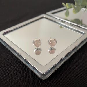Puede incluir: Un par de pendientes de botón redondos con piedras preciosas de color rosa pálido engastadas en plata. Los pendientes se reflejan en un espejo compacto plateado, sobre un fondo negro.
