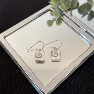 May include: A pair of dangle earrings featuring square-shaped white shell components with a natural brown spiral pattern. Each earring is set in a silver-toned metal frame and attached to a curved silver-toned ear wire for hanging support.