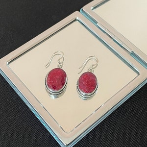 May include: A pair of silver earrings with red oval-shaped stones. The earrings are hanging from a mirror surface.