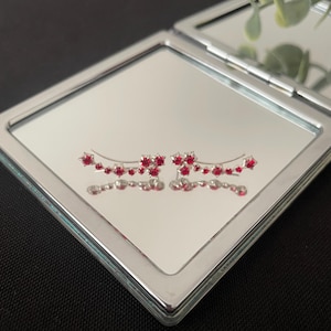 May include: A pair of silver-toned ear climbers with red gemstone accents, arranged in a floral design. The earrings are displayed on a mirrored surface within a silver compact, reflecting the design. The background is a dark surface.