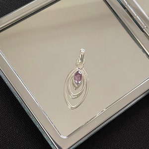 以下が含まれることがあります： 中央に紫色の宝石がついたシルバーのペンダント。ペンダントは、同心円状に3つの円が描かれた楕円形です。