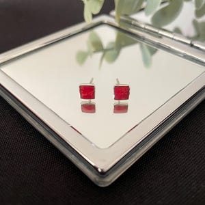 May include: A pair of square stud earrings featuring vibrant red stone centres set in polished silver-toned metal frames. The earrings are positioned on a reflective square surface, creating a clear mirror image of the red square studs below.