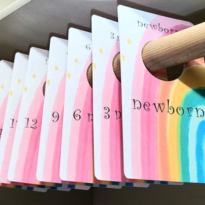 May include: A set of 10 pink and white rainbow-themed closet dividers with gold stars and black numbers. The dividers are labeled with the following sizes: newborn, 3 months, 6 months, 9 months, 12 months, 18 months, 24 months, 1, 2, and 3.