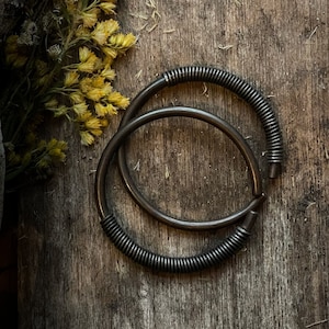May include: Two silver hoops with a textured, coiled design. The hoops are stacked on top of each other and are lying on a wooden surface.