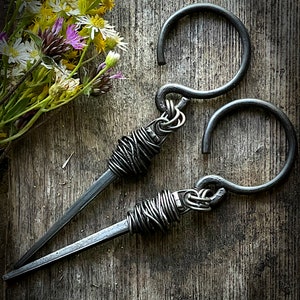 May include: Two black metal hooks with a twisted wire design. The hooks are on a wooden surface.