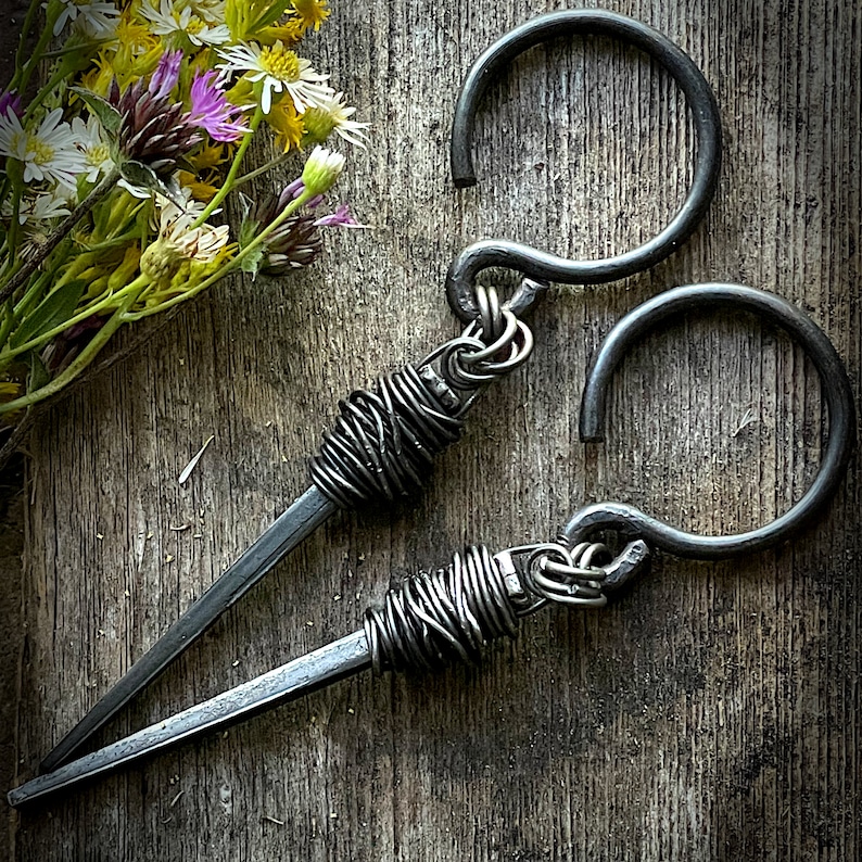 May include: Two black metal hooks with a twisted wire design. The hooks are on a wooden surface.
