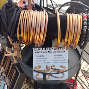 Könnte beinhalten: Kupferarmbänder und Perlenarmbänder auf einem schwarzen Ständer. Die Kupferarmbänder sind in verschiedenen Kupfer- und Schwarztönen gehalten. Ein Schild zeigt "Vorteile von Kupfer".