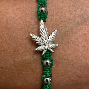 May include: Green and silver bracelet with a silver marijuana leaf charm. The bracelet is made of green string and silver beads.