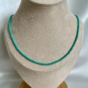 May include: A delicate turquoise beaded necklace displayed on a mannequin bust. The necklace is a single strand of small, evenly spaced beads, creating a simple yet elegant accessory. The beads are a vibrant shade of turquoise.
