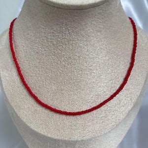 May include: A delicate red beaded necklace displayed on a mannequin. The necklace is composed of small, evenly spaced beads, creating a continuous strand. The vibrant red color of the beads contrasts with the neutral background, highlighting the jewelry's simple elegance.