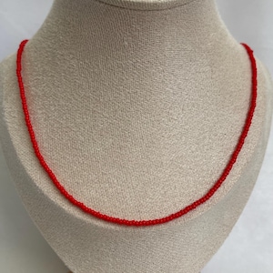 May include: A delicate red beaded necklace displayed on a mannequin. The necklace is composed of small, evenly spaced beads, creating a continuous strand. The vibrant red color of the beads provides a striking contrast against the neutral background.