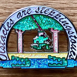 May include: A glittery enamel pin with a green pixelated figure swinging from a rope between two trees. The pin is shaped like a circle with the text "Obstacles are stepping stones" written around the edge.