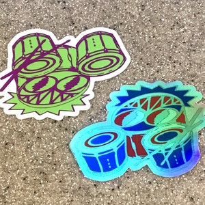 May include: Two colorful stickers featuring a drum set with drumsticks. The first sticker is green and yellow with a cartoon face and the second sticker is blue and red with a holographic finish.
