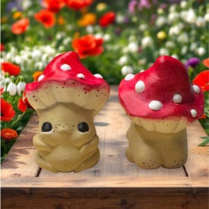 Può includere: Due fantasiose statuette a forma di fungo con cappelli rossi e pois bianchi. I gambi dei funghi sono beige con dettagli a pois neri. Le statuette sono su una superficie di legno con uno sfondo floreale sfocato.