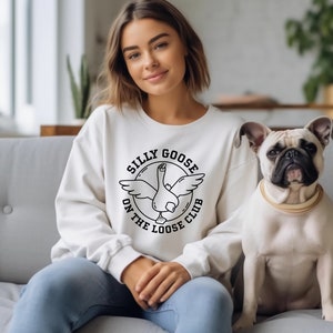 Puede incluir: Una sudadera blanca con un gráfico negro de un ganso con el texto "Silly Goose On The Loose Club". Una persona lleva la sudadera y está sentada en un sofá con un perro.