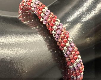 May include: A beaded bracelet with a spiral design. The bracelet features a mix of red, pink, and silver beads. The beads are tightly woven together, creating a textured surface. The bracelet is shown on a dark surface.