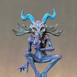 May include: A small, painted figurine of a creature with a skull-like face, blue horns, and a green and purple body. The creature is sitting on a decorative pillar with a round top.