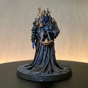 May include: Miniature figurine of a robed figure with a crown and bells, holding a scale with a small candle. The figure has blue skin and is painted in dark colors. The base is dark gray.
