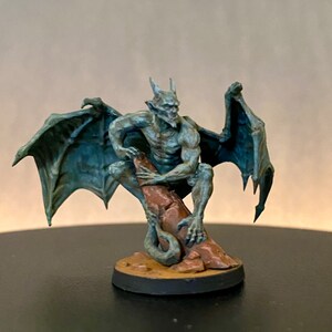 May include: A painted miniature figurine of a gargoyle with gray wings and a stone-like body, perched on a rock. The gargoyle has a menacing expression and sharp claws.