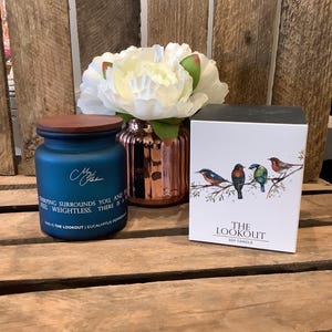 May include: A blue soy candle with a wooden lid and the text "Chirping surrounds you and feel weightless." A white box with a bird illustration and the text "The Lookout Soy Candle" is next to it. A white flower and a copper vase are also in the image.