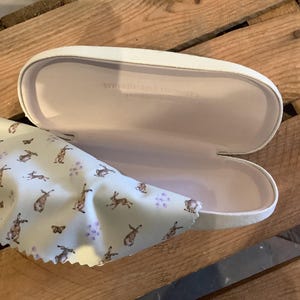 May include: An open white eyeglasses case with a light pink interior and the text "WEIDMANNS DESIGN". A light blue cleaning cloth with a pattern of brown rabbits and purple flowers is next to the case.