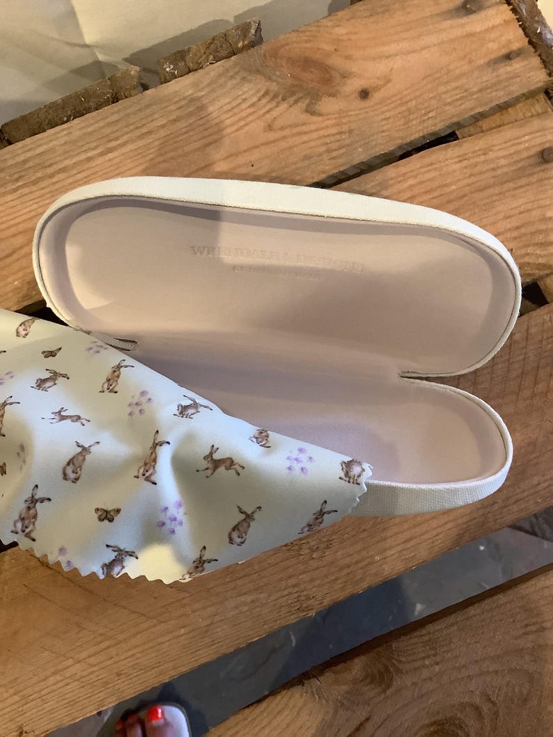 May include: An open white eyeglasses case with a light pink interior and the text "WEIDMANNS DESIGN". A light blue cleaning cloth with a pattern of brown rabbits and purple flowers is next to the case.